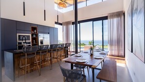 A modern kitchen with a central island, bar stools, and a dining area with a table set for two. - Oceanvista Villa I Santa Teresa Beach  Brand New Oceanview Villa Gated Community (santa teresa)