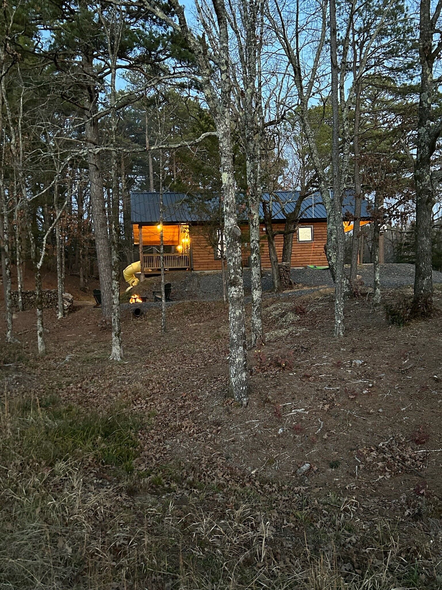 Una cabaña rodeada de árboles, con un porche visible y un pozo de fuego.