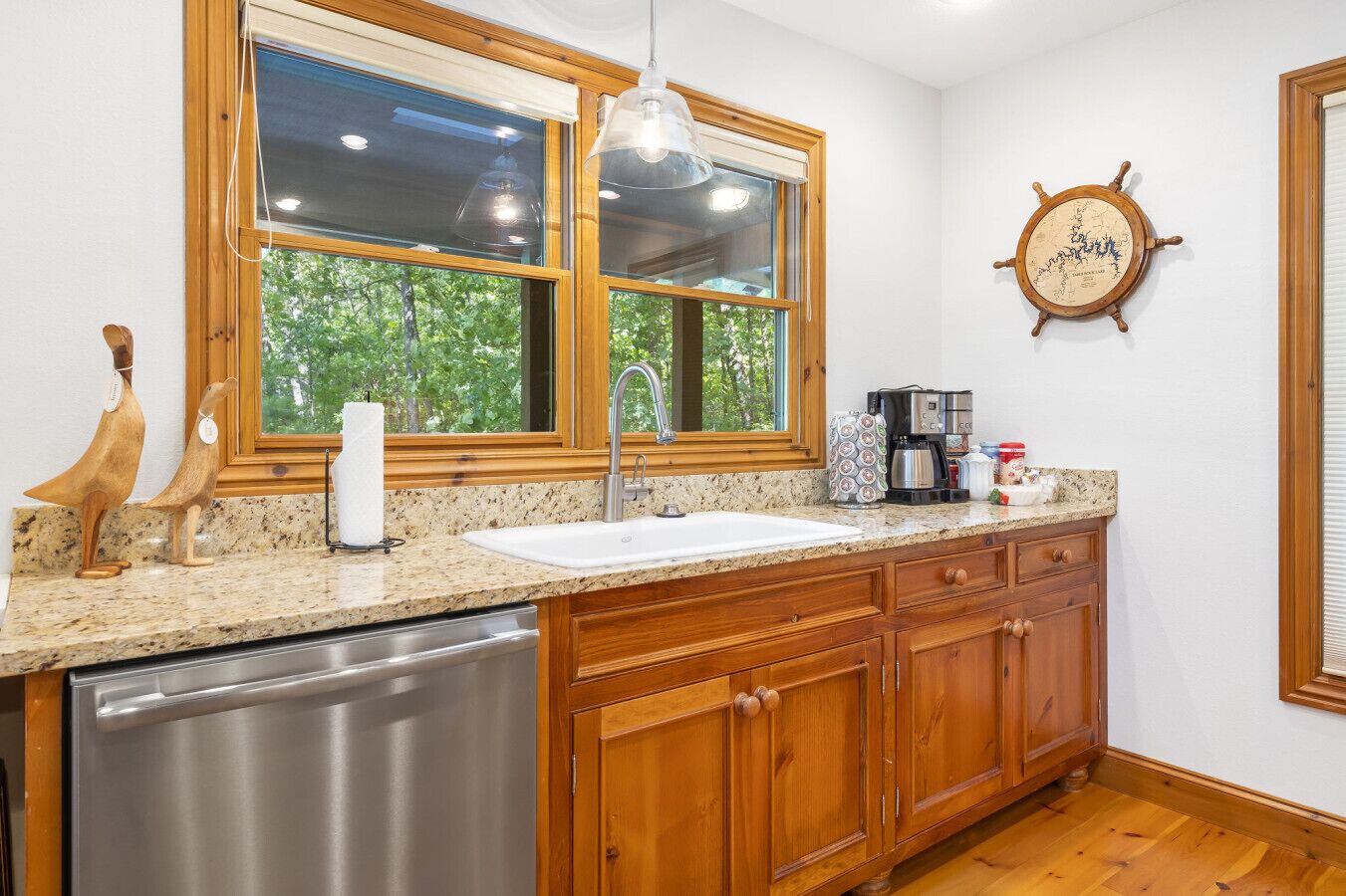A kitchen with wooden cabinets, a stainless steel dishwasher, a double sink, and a window with a view of trees.
