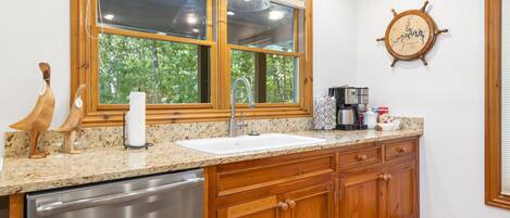 A kitchen with wooden cabinets, a stainless steel dishwasher, a double sink, and a window with a view of trees.