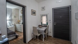 Een knus vertrek met een eettafel, stoelen, een donkere deur, een klein raam met kantgordijnen en een klok aan de muur.