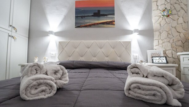 Een opgemaakt bed met een grijze deken, twee opgerolde handdoeken en een schilderij van een zonsondergang aan de muur.