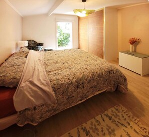 Quarto com cama, mesa de cabeceira, janela e vista para o verde lá fora.