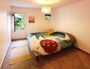 Un dormitorio con cama, una puerta, una ventana, una lámpara colgante y un pequeño adorno en la pared.