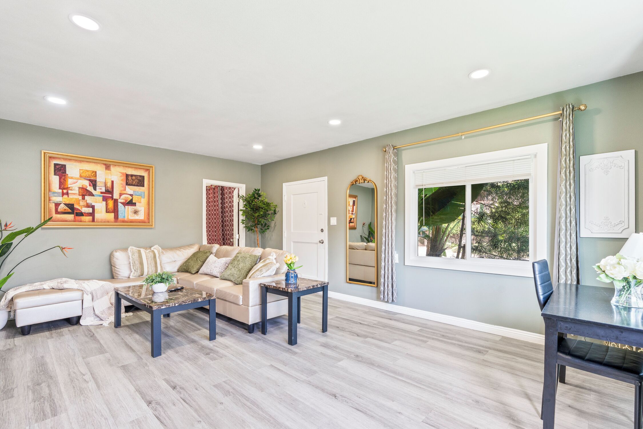 A spacious living room with a sofa set, a coffee table, and a dining table. There is a large window with curtains, a mirror, and a painting on the wall.