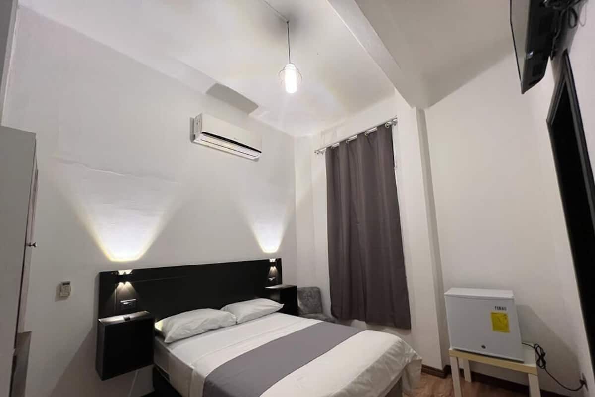 A hotel room with a bed, bedside tables, an air conditioning unit, a TV, and a small desk with a power outlet.