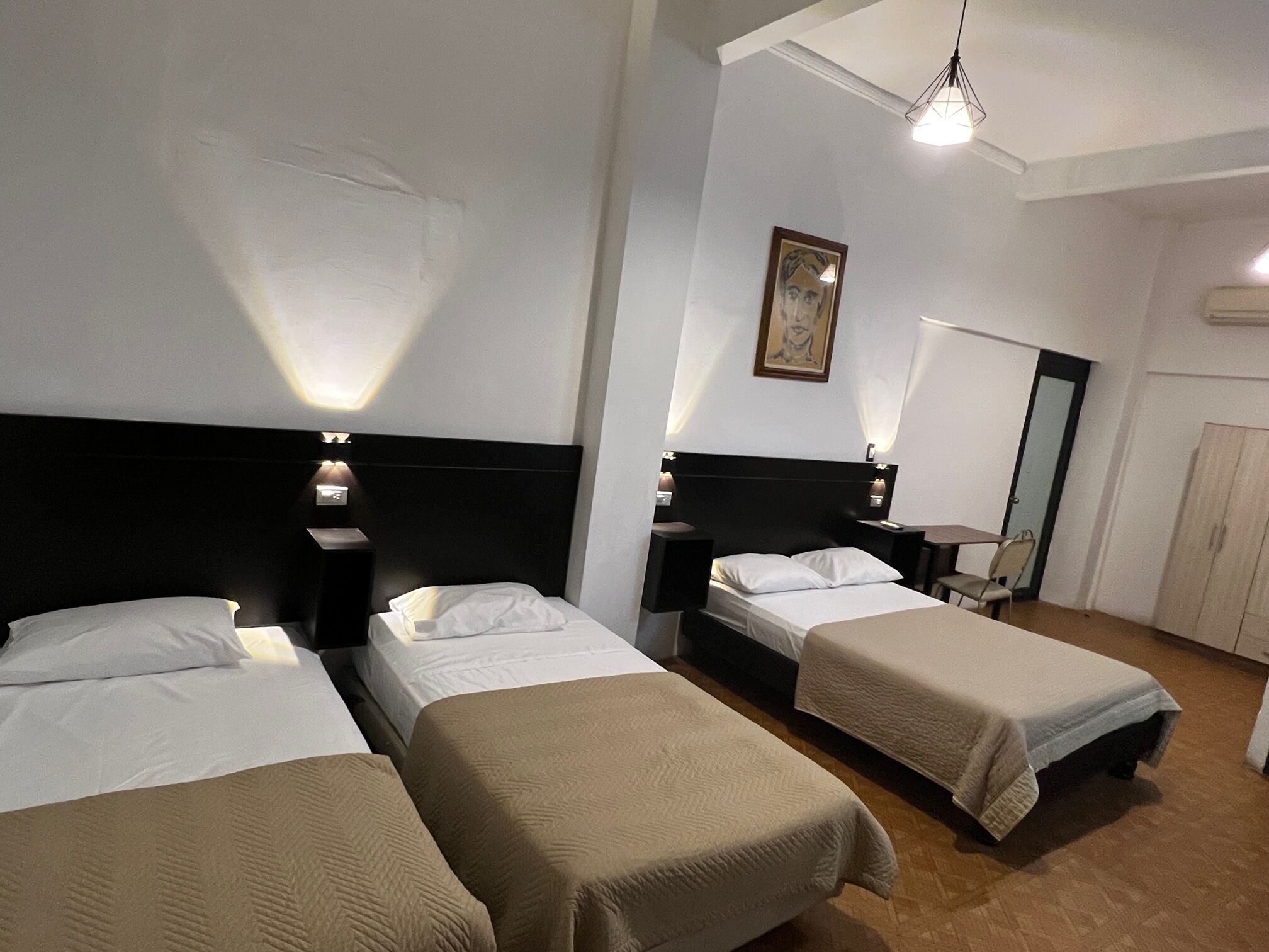 Une chambre d’hôtel avec trois lits simples, chacun doté d’une tête de lit et de tables de chevet. Un tableau orne le mur et une armoire se trouve dans un coin.
