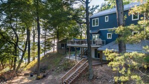 A two-story house with a deck, surrounded by trees and a clear path leading to the entrance. - Tranquil Shores | Lake Michigan | Private Beach | Sleeps 11 (Norton Shores)
