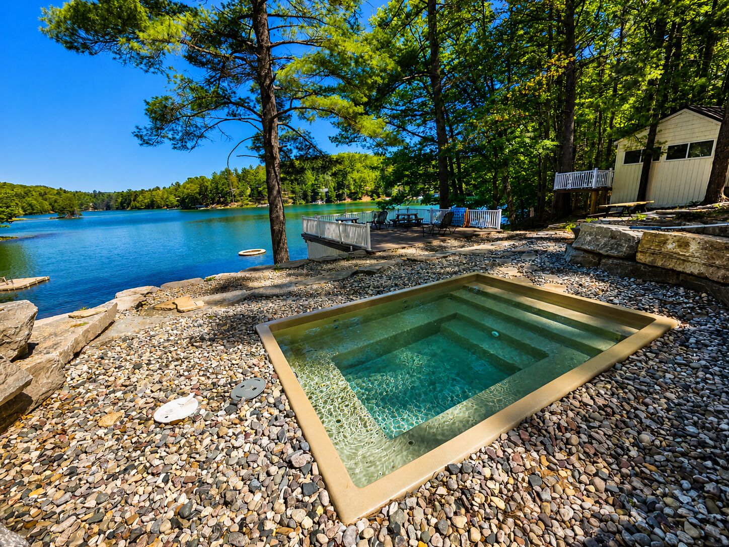 Una piscina con borde de guijarros, muelle de madera y vista al lago.