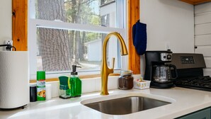 Una cocina con un lavamanos de acero inoxidable, un grifo dorado, una cafetera y una ventana con vistas a árboles.