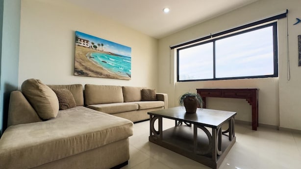 Una sala de estar con un sofá color beige, una mesita de centro y un cuadro que muestra una escena de playa.