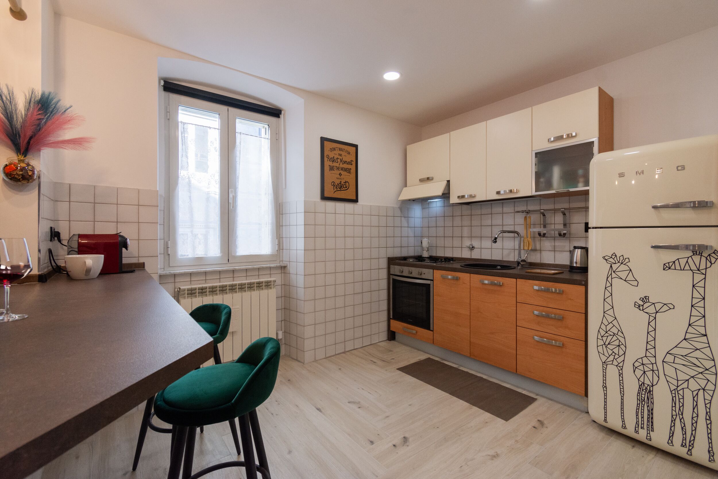 A kitchen with a wooden countertop, two green bar stools, a refrigerator with giraffe designs, and a window with a view.