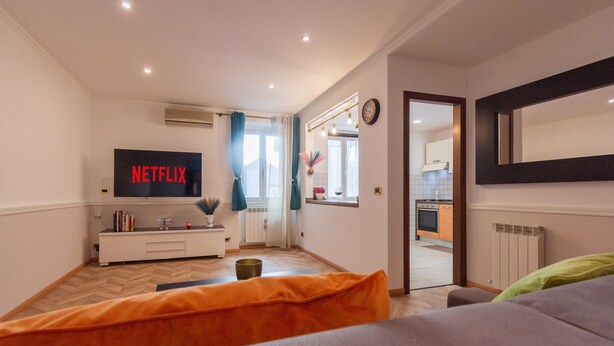 A modern living room with a flat-screen TV displaying 'NETFLIX', a sofa, a coffee table, and a kitchen visible through an open door.