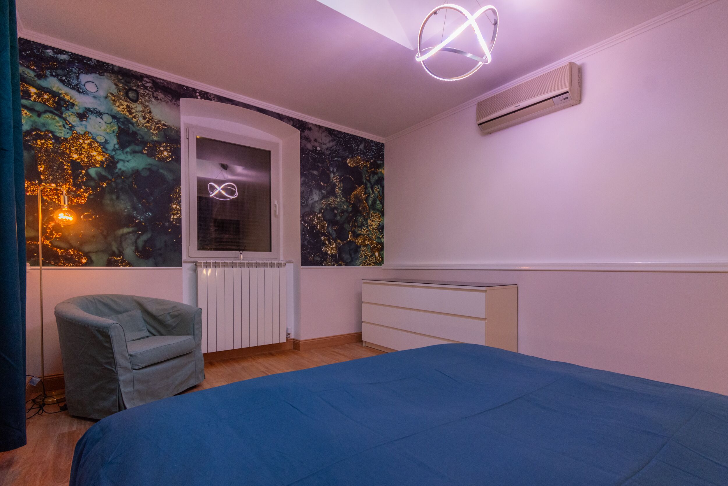 Un dormitorio con una cama, una silla, un radiador, aire acondicionado instalado en la pared y una lámpara en el techo.