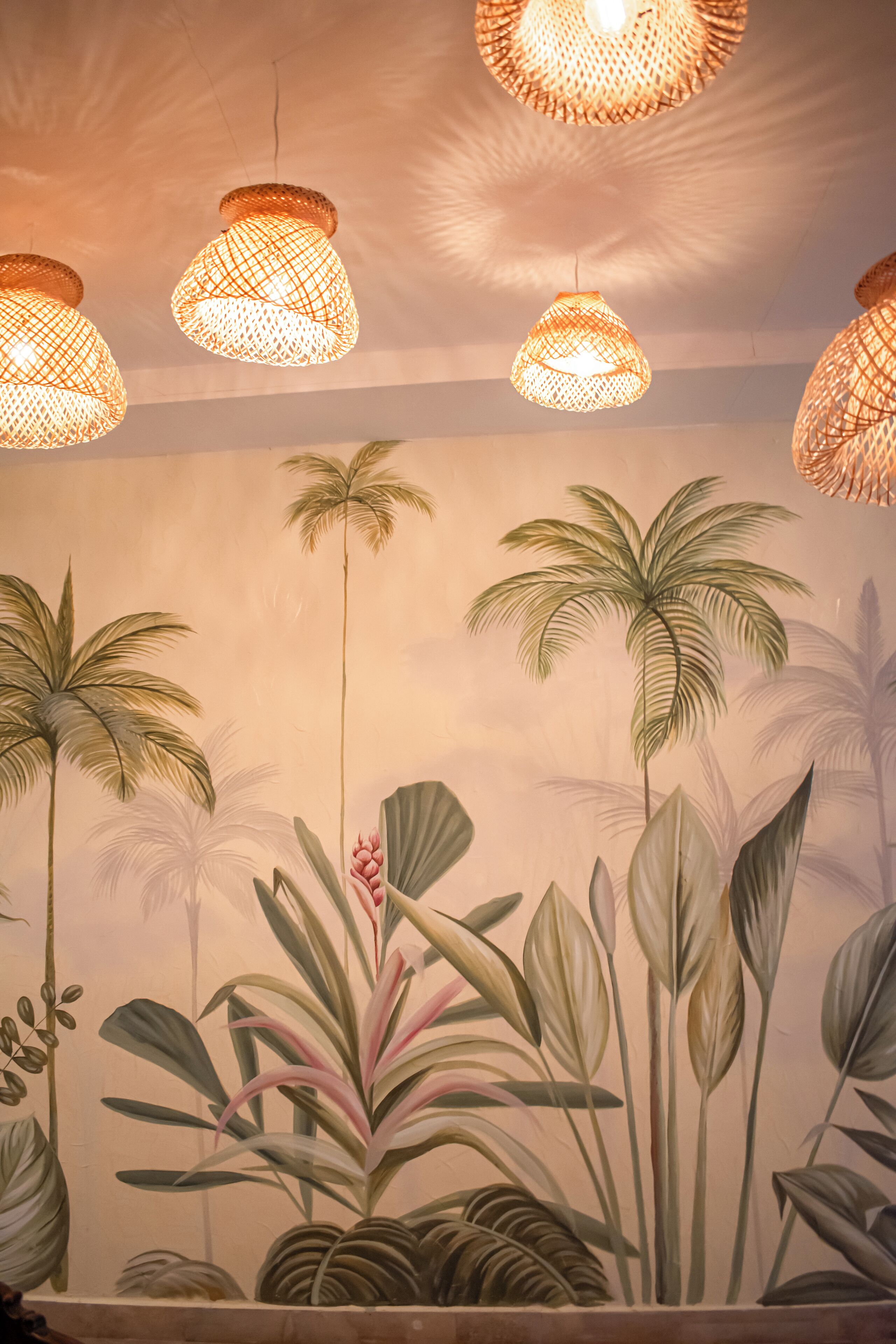Een kamer met een tropische muurschildering, hangende geweven lampen en een groot raam met uitzicht op palmbomen.