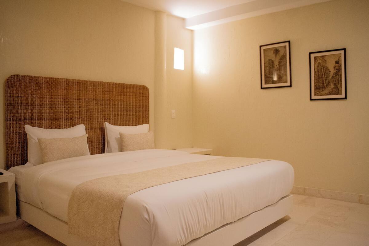 Una cama bien hecha con cabecera, una mesita de noche y dos cuadros enmarcados en la pared.