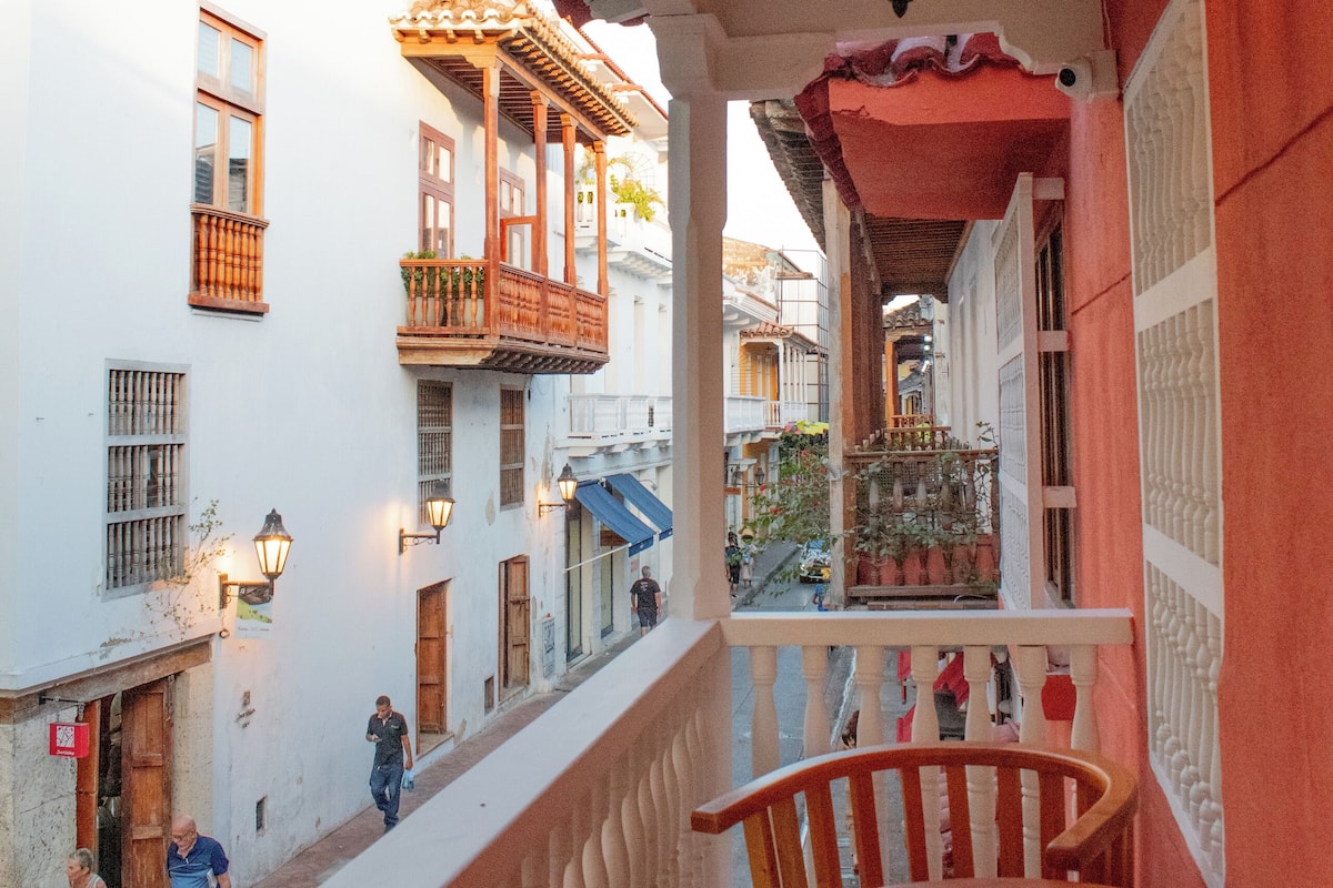 Un balcón con una mesa y sillas de madera, con vista a una calle con edificios y un farol colgante.