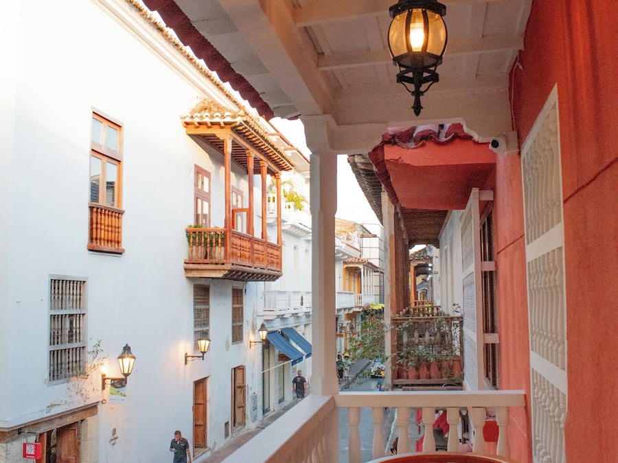 A balcony with a wooden table and chairs, overlooking a street with buildings and a hanging lantern.