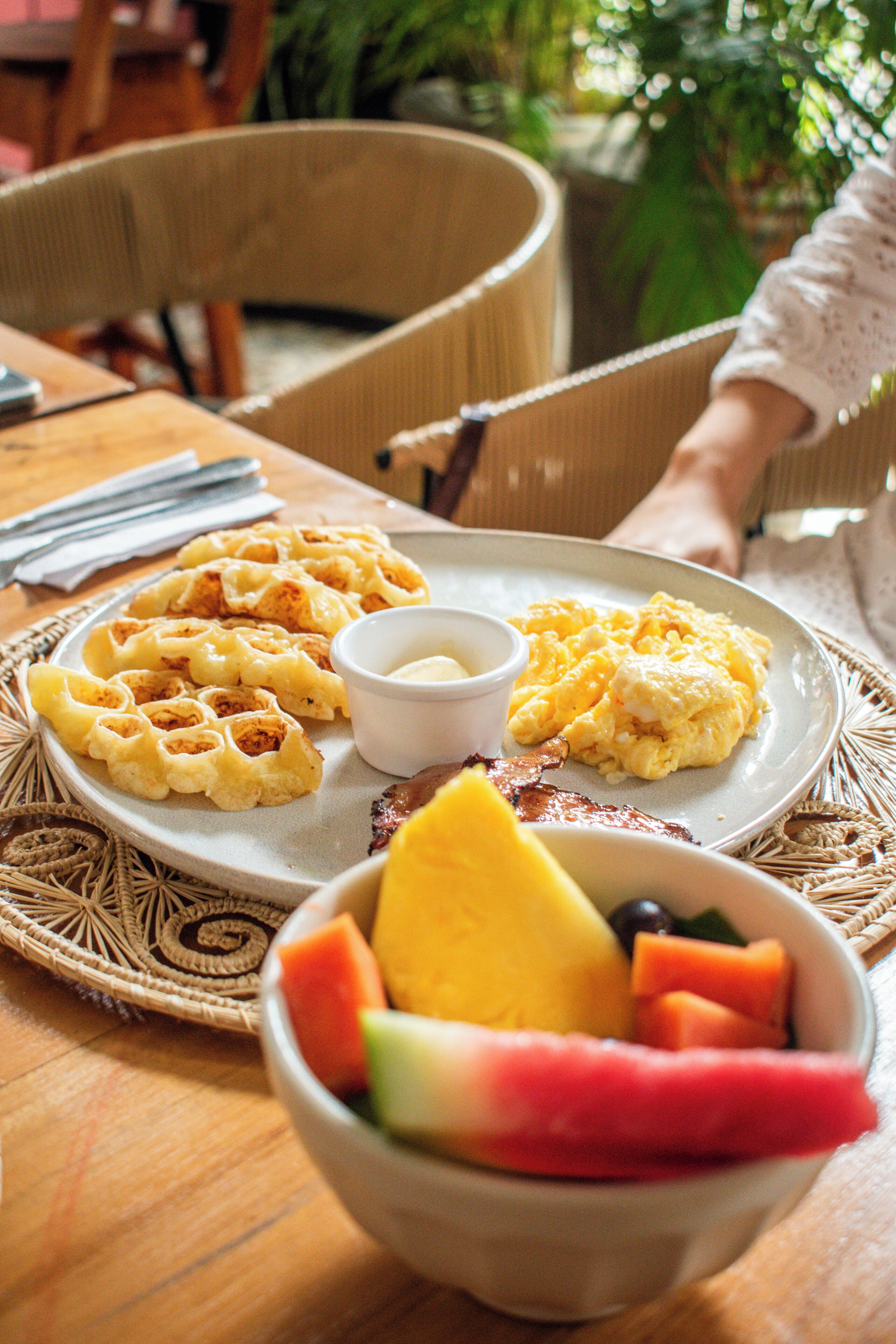 A breakfast plate with waffles, scrambled eggs, and bacon, accompanied by a bowl of fruit.