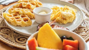 Un plato de desayuno con waffles, huevos revueltos y tocino, acompañado de un bol de fruta.