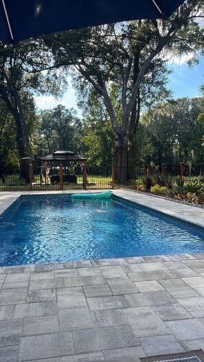 A rectangular swimming pool with a tiled deck, surrounded by a fence and trees. - Tiny House on The Grove (La Crosse)