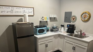 Una cocina con refrigerador, microondas, cafetera y pileta. Hay un cartel en la pared y un reloj también.