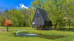 Eine rustikale Holzhütte mit Metalldach, auf Stelzen stehend, umgeben von üppigem Grün und einem kleinen Teich.