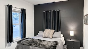 A bedroom with a bed, a nightstand, a window with curtains, and a view of the outside. - The Bunga Glow: Modern, Cozy & Bright Near Downtown (Sioux Falls)