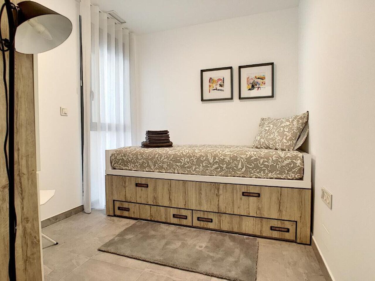 A bedroom with a bed, a bedside lamp, a small bedside table, and a window with curtains.