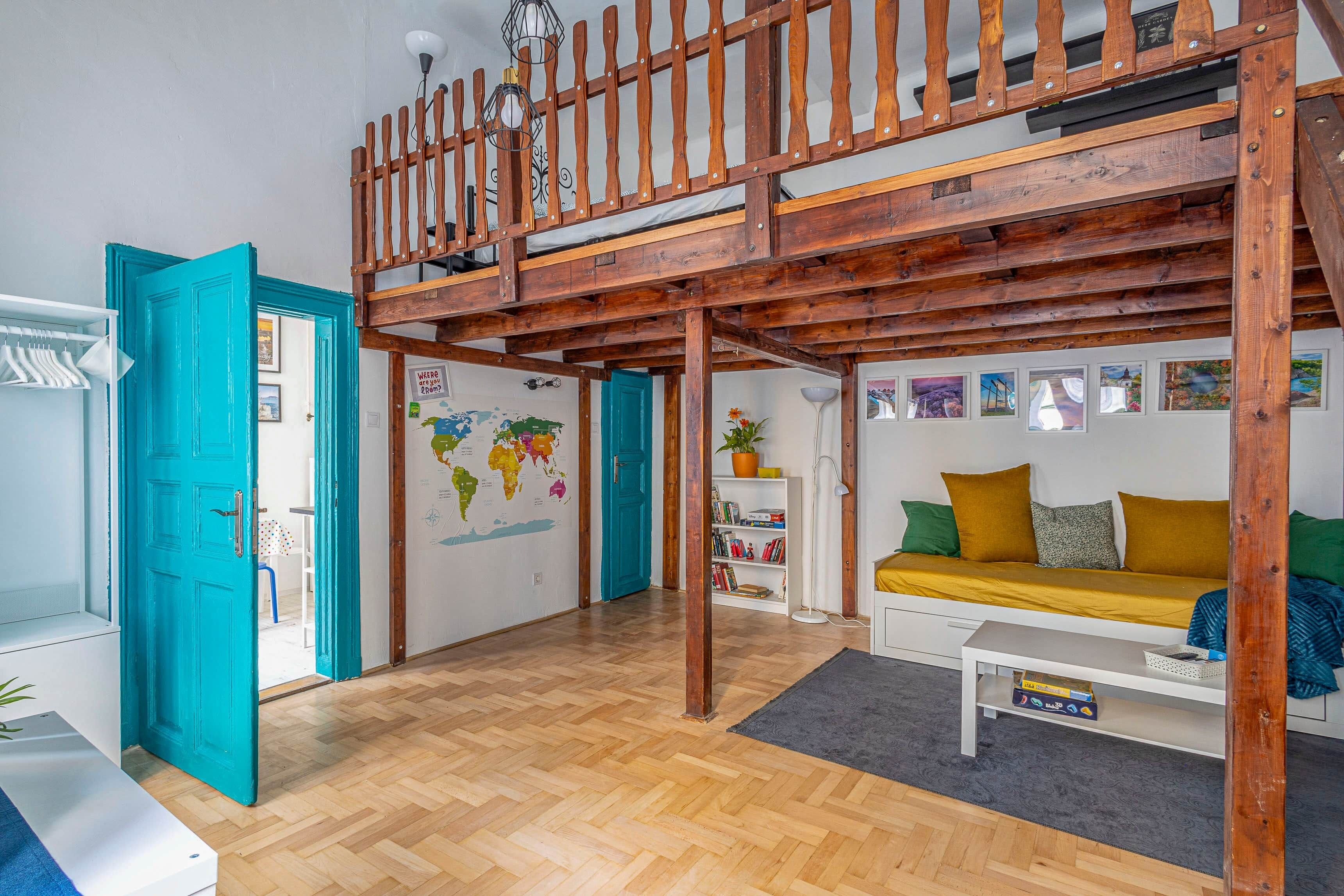 A loft-style room with a wooden ceiling, a bunk bed, a world map on the wall, a sofa with cushions, and a bookshelf.