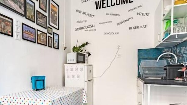 Una stanza con una parete bianca decorata da insegne bilingue con la scritta 'WELCOME', un tavolino con tovaglia a pois, una sedia blu e un armadietto bianco con una pianta sopra.