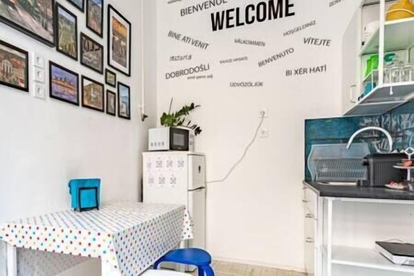 A room with a white wall decorated with multilingual 'WELCOME' signs, a small table with a polka dot tablecloth, a blue chair, and a white cabinet with a plant on top.