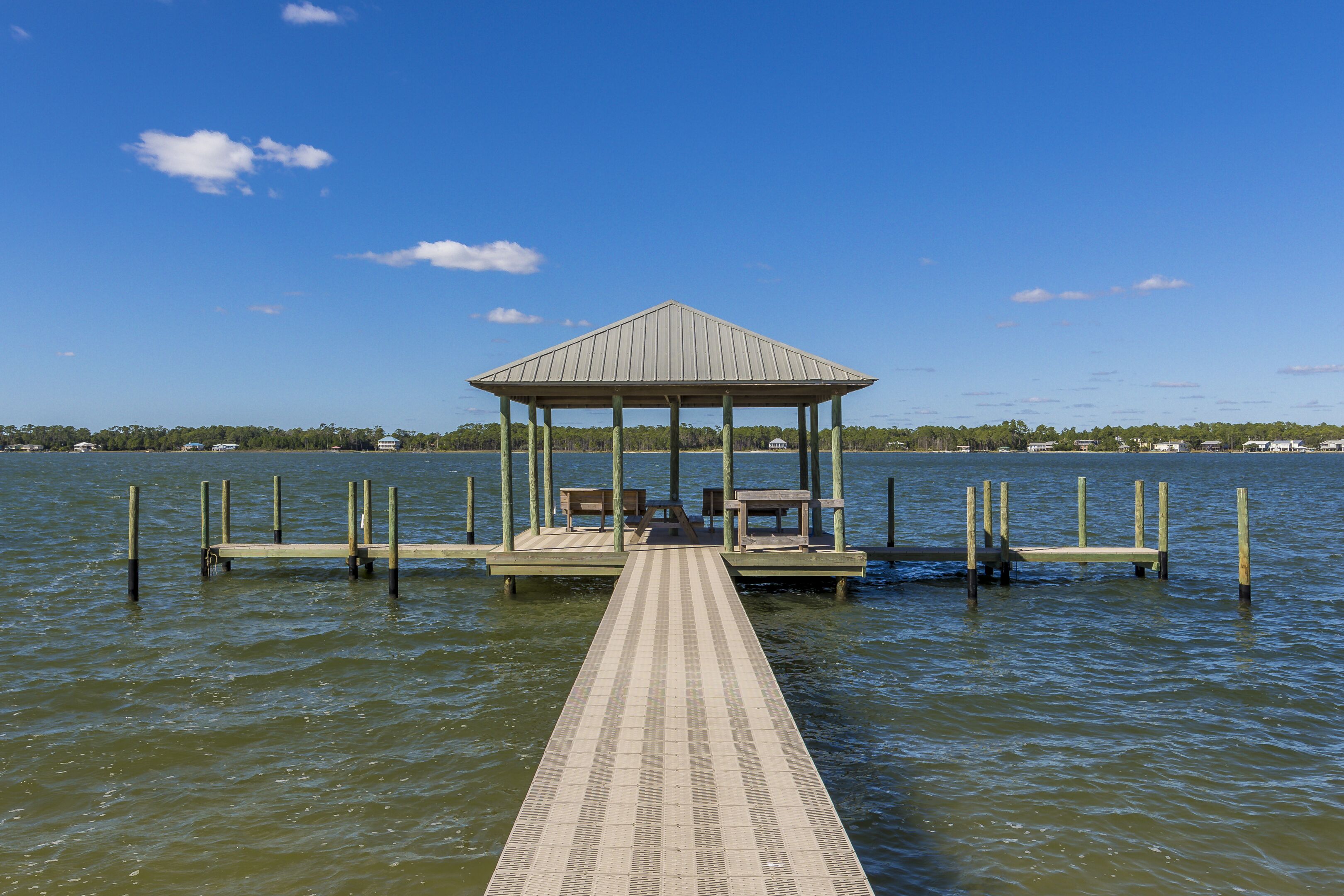 A wooden pier leading to a gazebo with benches, surrounded by calm water and a clear sky.