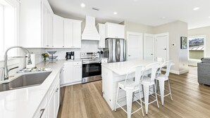 A modern kitchen with white cabinetry, stainless steel appliances, and a central island with bar stools. - Amazing New Lagoon View Home (Gulf Shores)