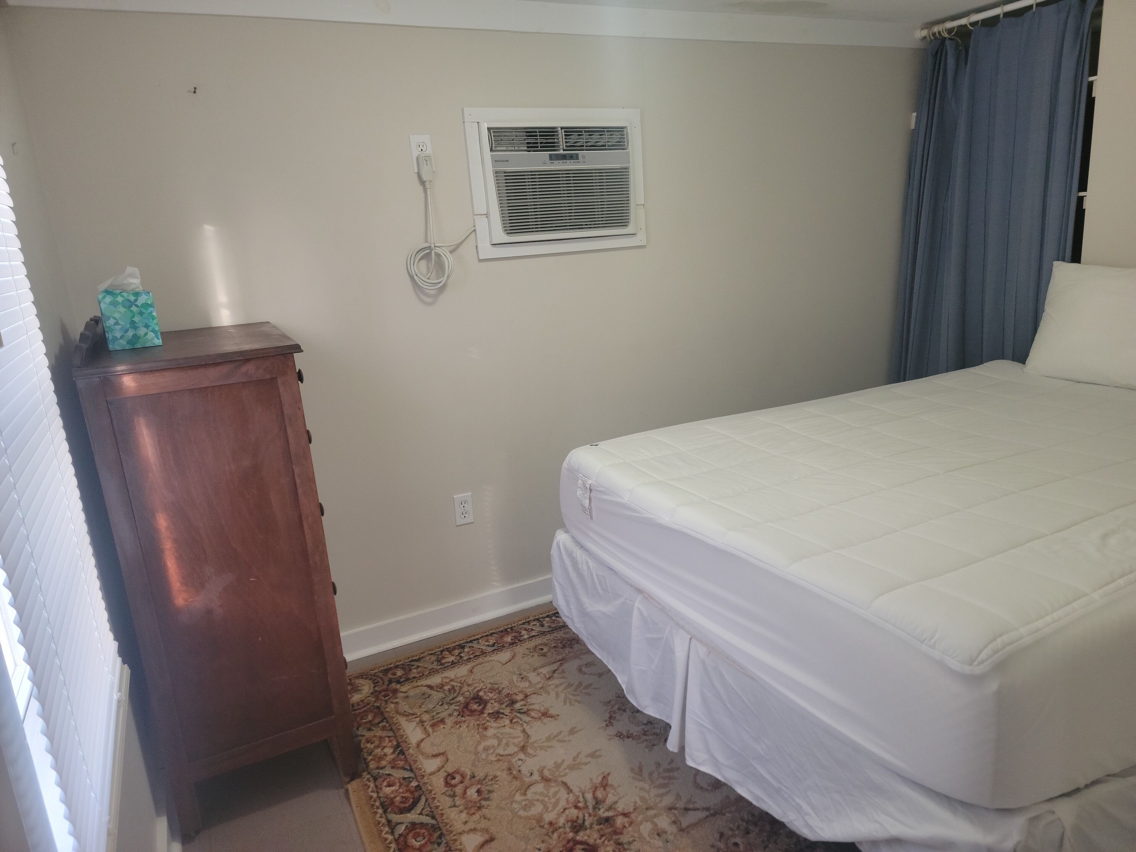 A bedroom with a bed, a wooden dresser, a wall-mounted air conditioner, and a patterned rug.