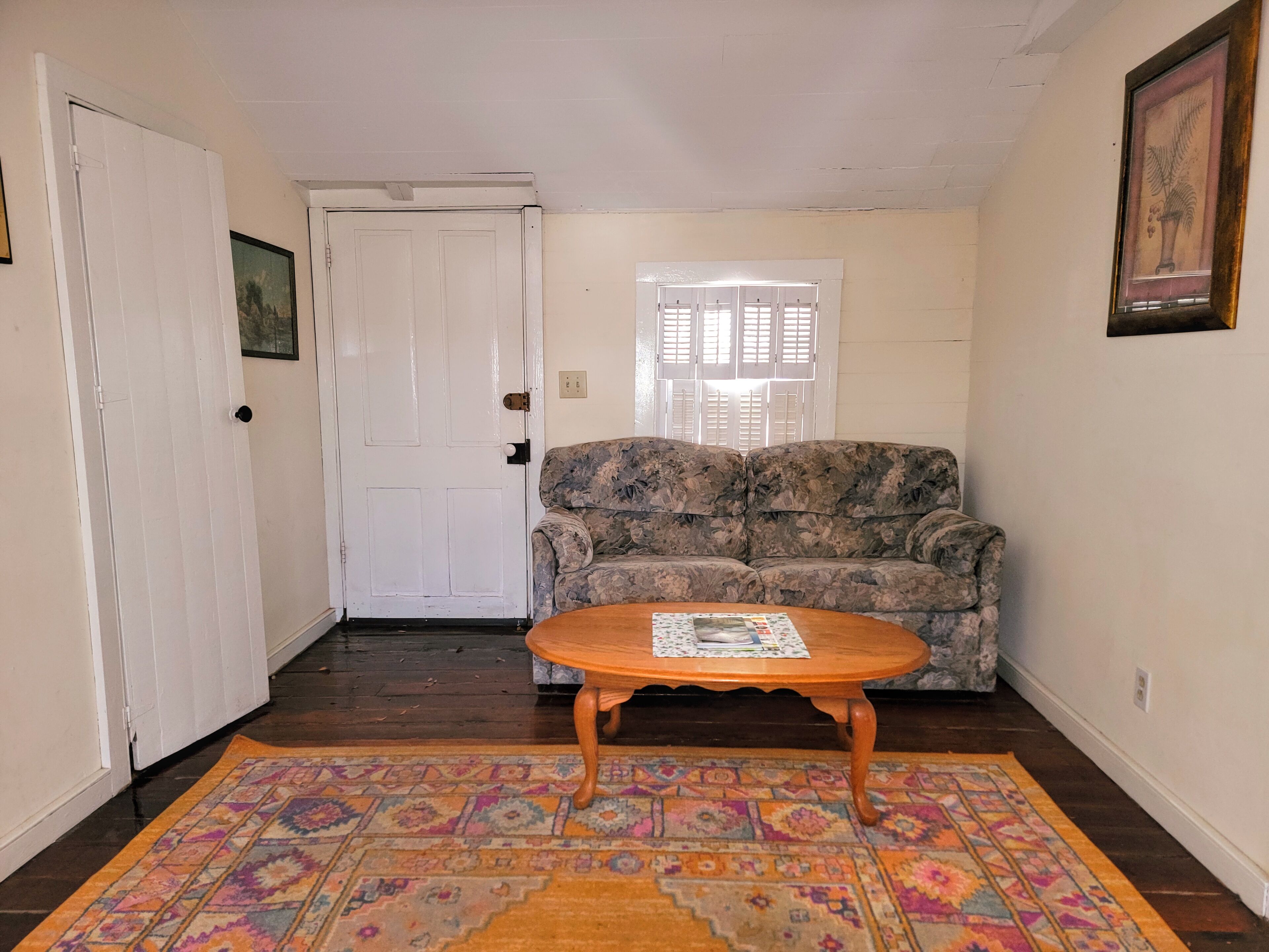 A room with a patterned rug, a wooden coffee table, a floral sofa, a white door, and a window with shutters.