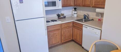 A compact kitchen with wooden cabinets, a white refrigerator, a microwave, a stove, a coffee maker, a sink, and a dishwasher.