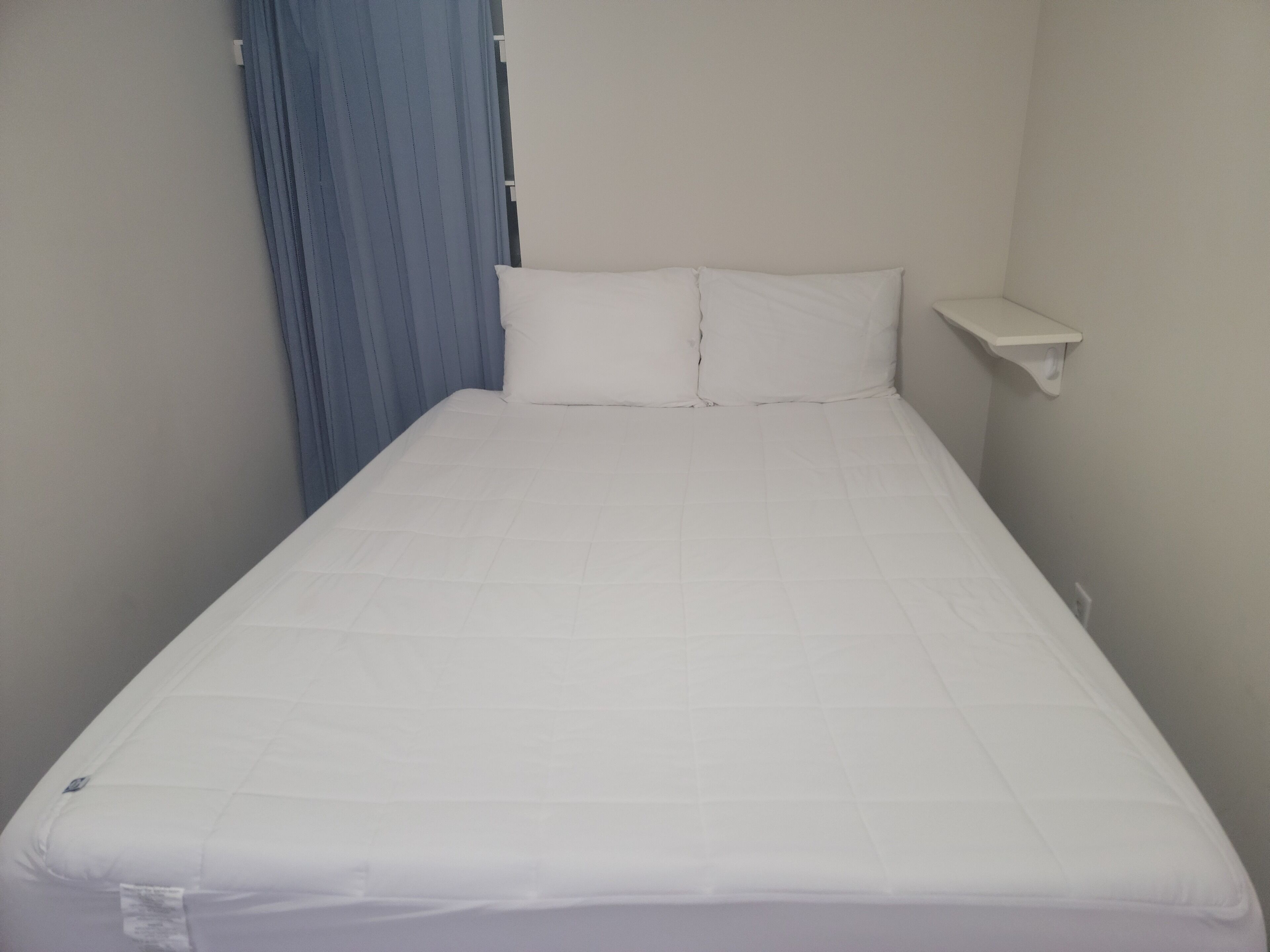 Quarto pequeno com uma cama de solteiro, colchão branco, cortina azul e mesa de cabeceira branca.