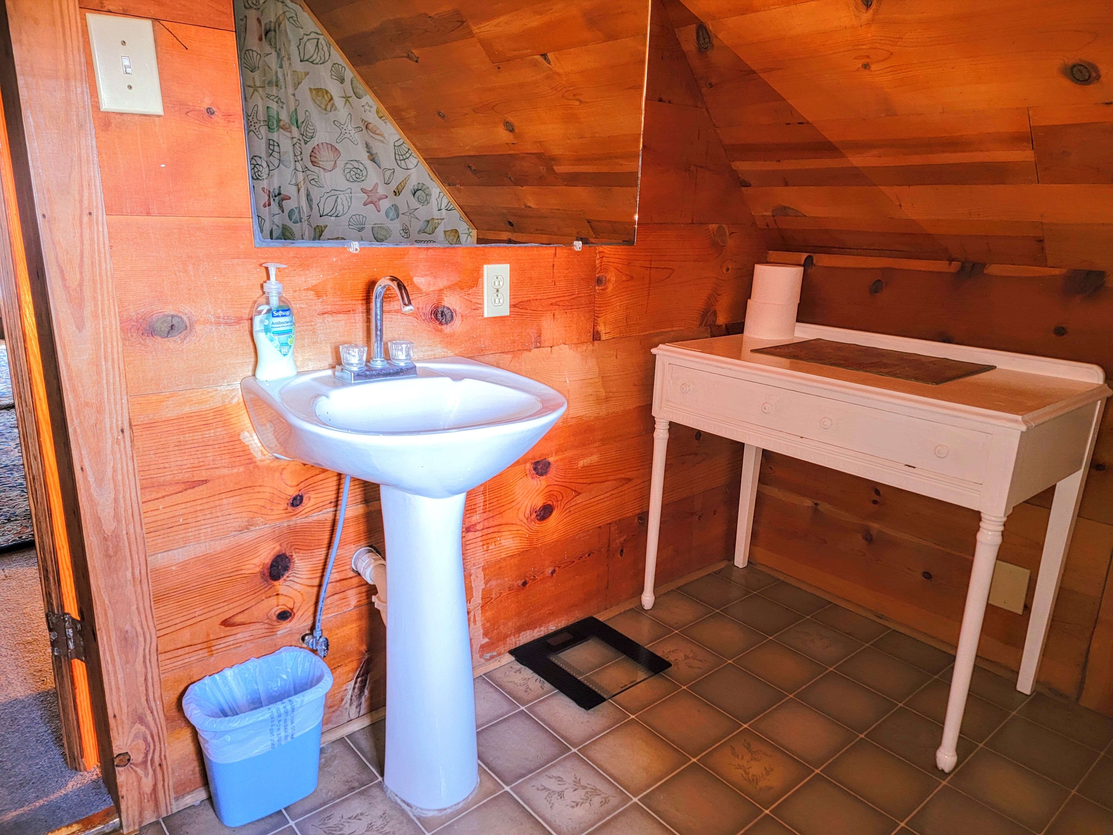 Un interior de madera con un lavabo blanco, un pequeño cubo de basura y una mesa blanca.
