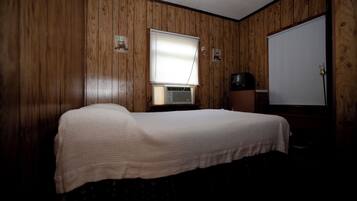 Een enkel bed in een kamer met houten lambrisering, een raam met jaloezieën, een televisie en een deur.