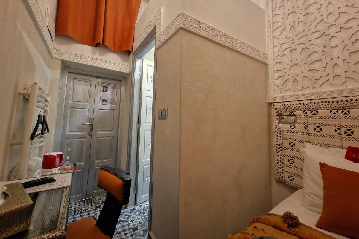 Habitación de hotel con cama, una mesita con una taza roja, una silla y una puerta que da a otra habitación.