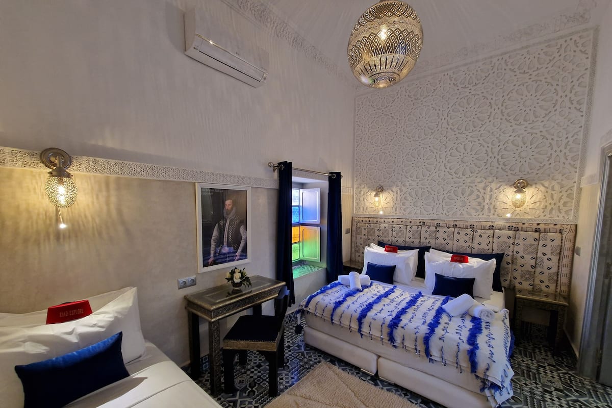 Habitación de hotel con cama, mesita de noche, espejo, aire acondicionado de pared, paneles decorativos y suelo estampado.