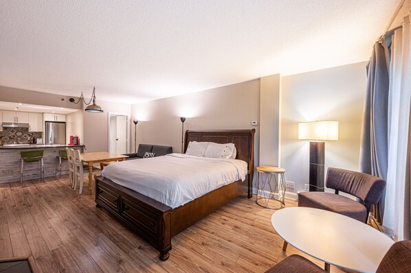 A hotel room with a wooden floor, a bed, a desk, a chair, a lamp, and a view of the kitchen.