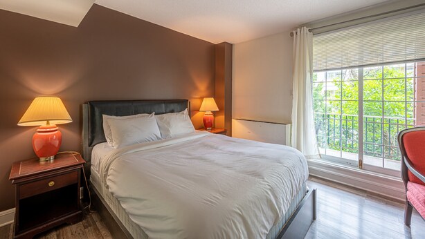 A hotel room with a large bed, two bedside lamps, a window with blinds, and a view of greenery outside.