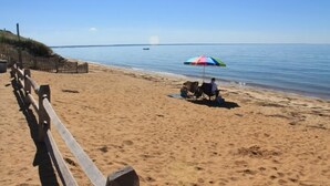 A sandy beach with a wooden fence, a colorful umbrella, and a person sitting on a chair.