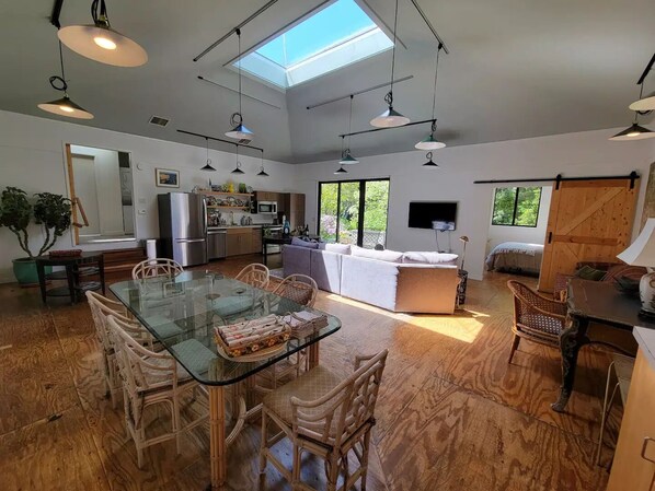 Una cocina moderna con una isla grande, comedor con mesa de vidrio y sillas de mimbre, y una sala de estar acogedora con sofá y televisión.