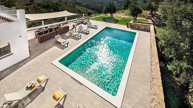 A modern outdoor swimming pool with lounge chairs and a dining area, surrounded by a landscaped garden and a view of distant hills.
