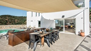 A poolside dining area with a long table and chairs, a pool, and a view of the surrounding landscape.