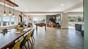 A spacious living area with a wooden dining table, colorful chairs, a foosball table, and a large window overlooking a scenic view.