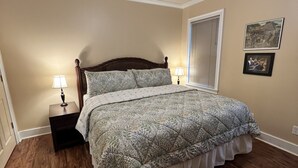 Un dormitorio con piso de madera, una cama con colcha estampada, dos lámparas de noche, un ventilador de techo y una ventana con persianas.